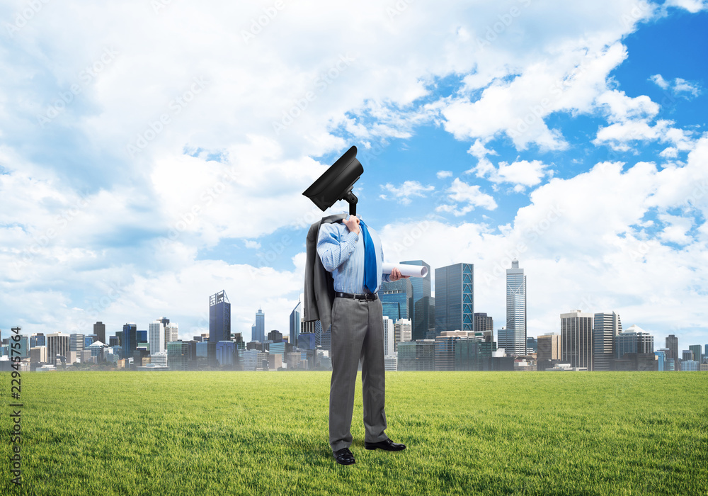 Camera headed man standing on green grass against modern cityscape