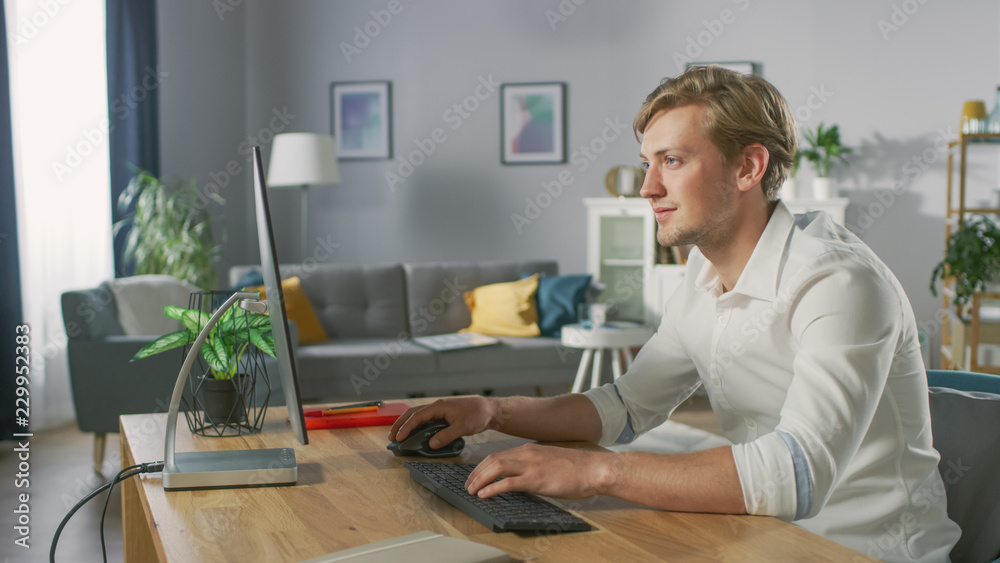 Handsome Young Designer Works On Personal Computer Sitting at His Desk. In the Background Stylish Ho