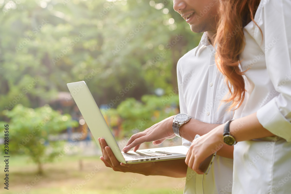 Man and woman in white shirt use laptop and smartphone together in outdoors park