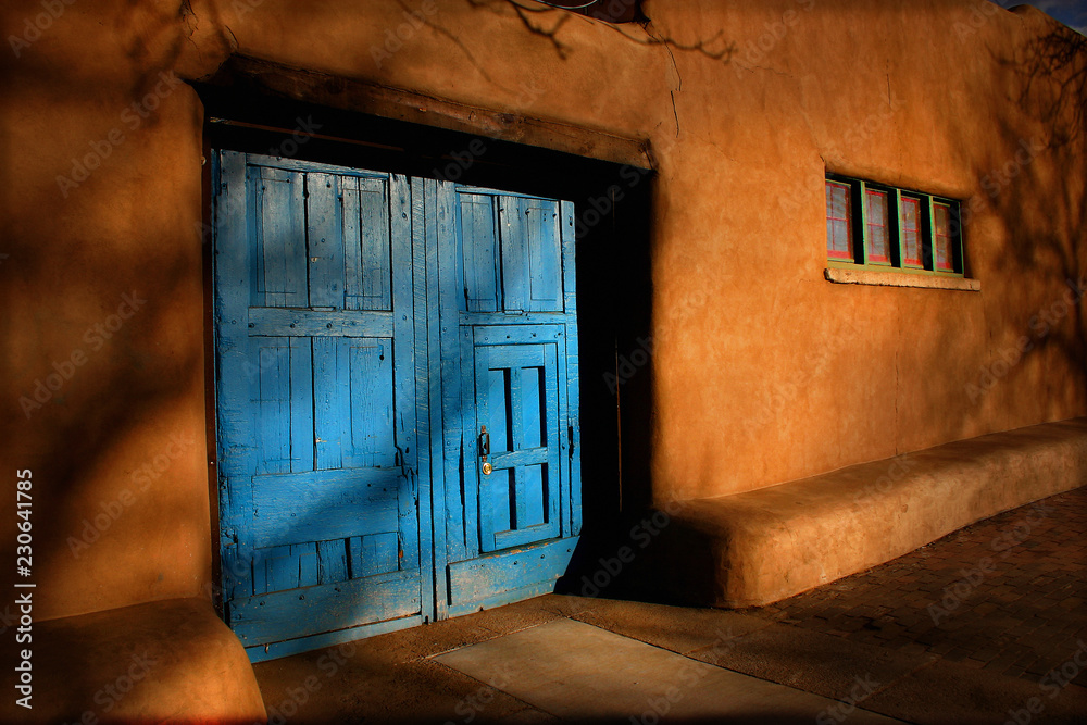 Bright blue wooden doors and Adobe walls of this Santa Fe, New Mexico street view with shadows of tr