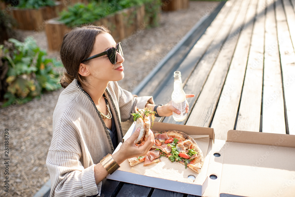 Young woman having a snack with pizza sitting outdoors at the modern public park