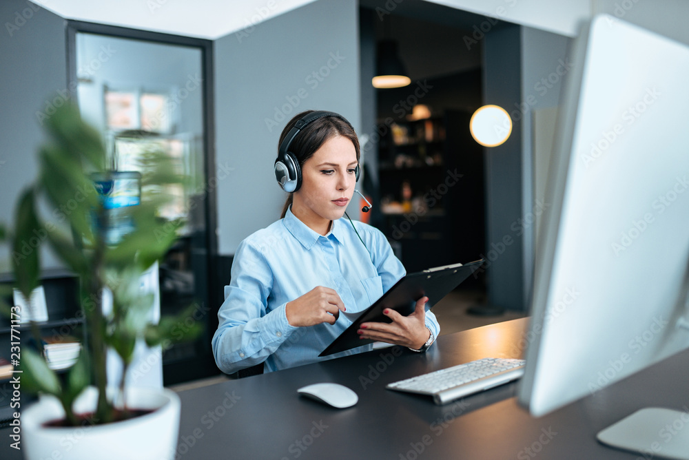 Young female help desk operator working with headset, computer and clipboard.