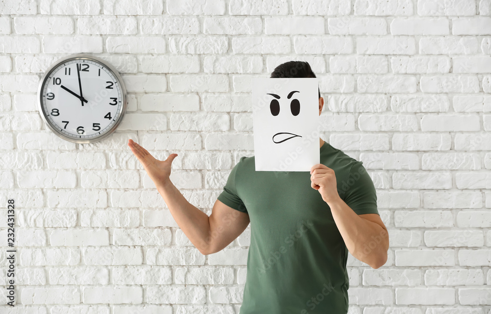 Young man hiding face behind sheet of paper with drawn emoticon against white brick wall with clock