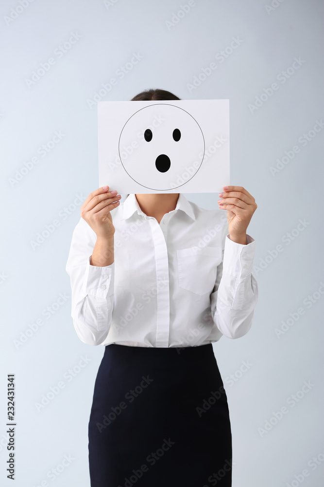 Young woman hiding face behind sheet of paper with drawn emoticon on light background