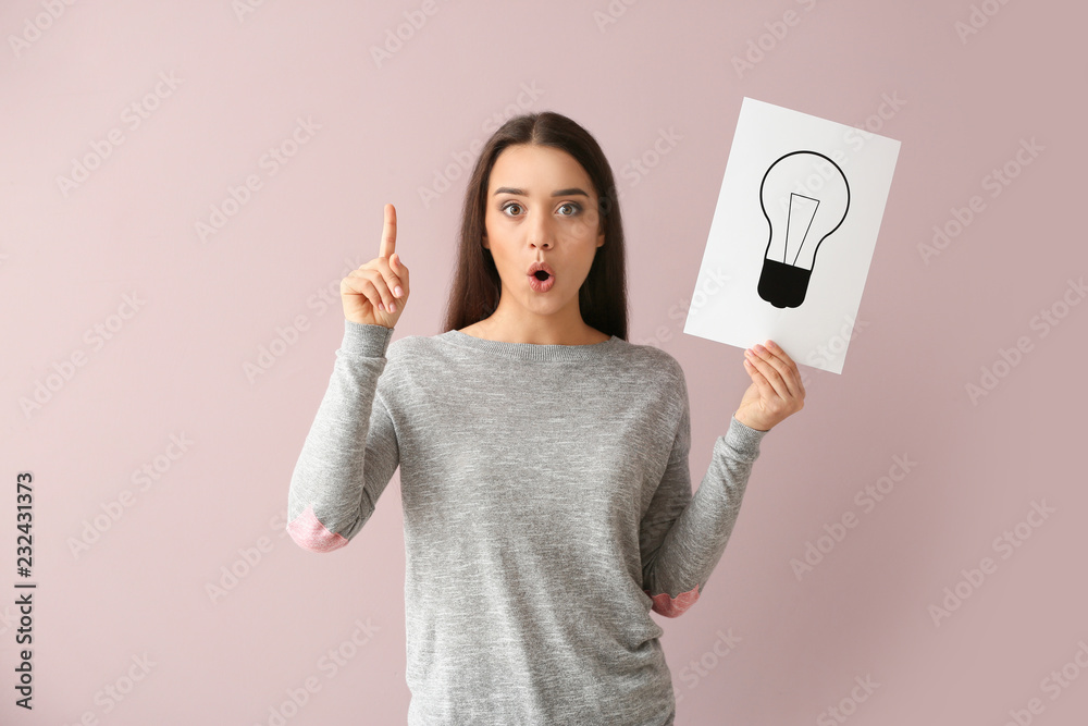 Emotional young woman holding paper sheet with drawn light bulb as symbol of idea on color backgroun