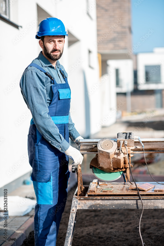 Builder in uniform cutting tiles with big electro saw on the construction site outdoors