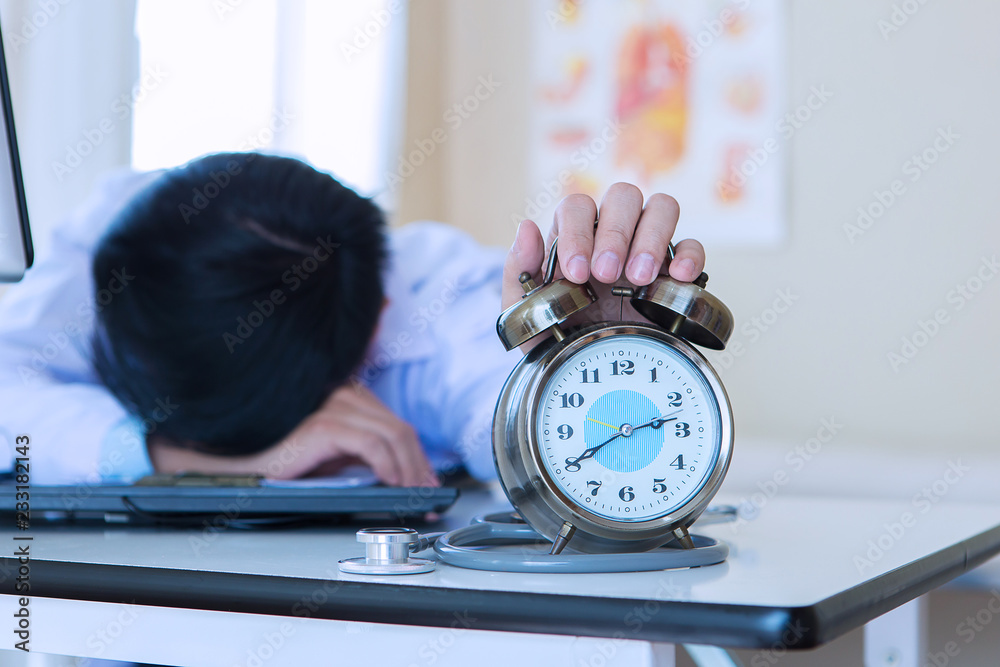 Sleepy, exhausted doctor holding alarm clock on desk in hospital