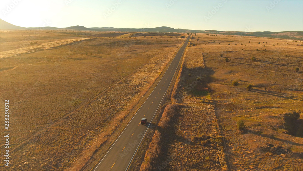 AERIAL: Flying above SUV car driving along empty countryside road at sunset