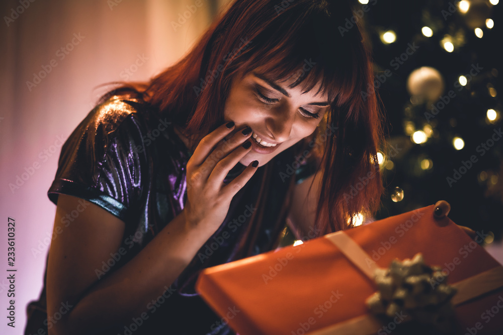 Delighted smiling woman opening Christmas gift box under tree at home