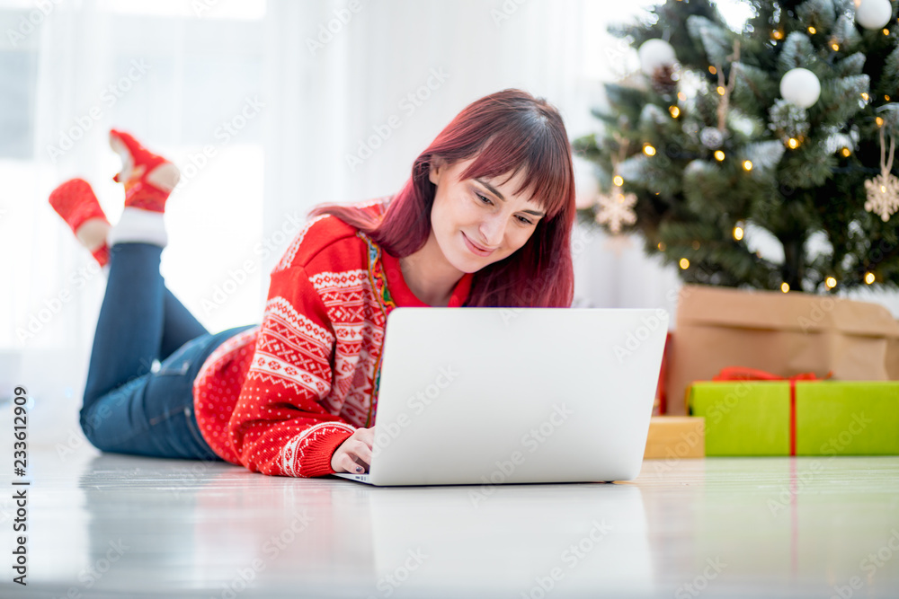 Smiling woman using laptop to Christmas shopping online lying on floor next to xmas tree