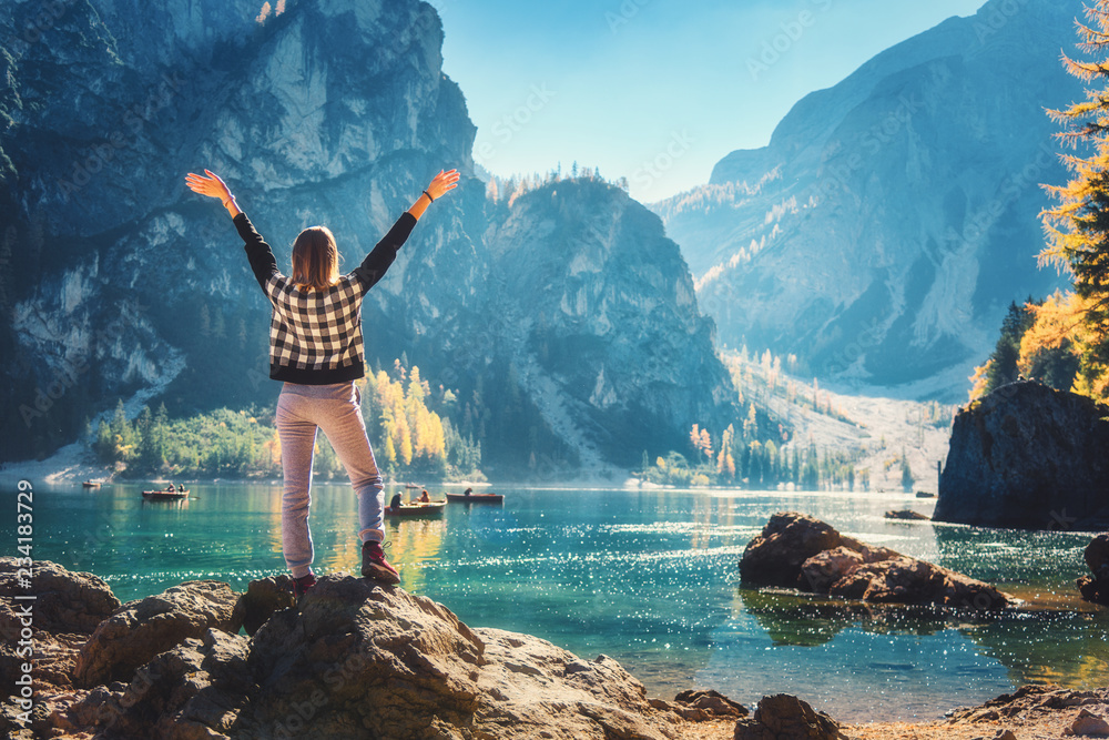 Standing woman on the stone with raised up arms on the coast of Braies lake at sunrise. Autumn in Do