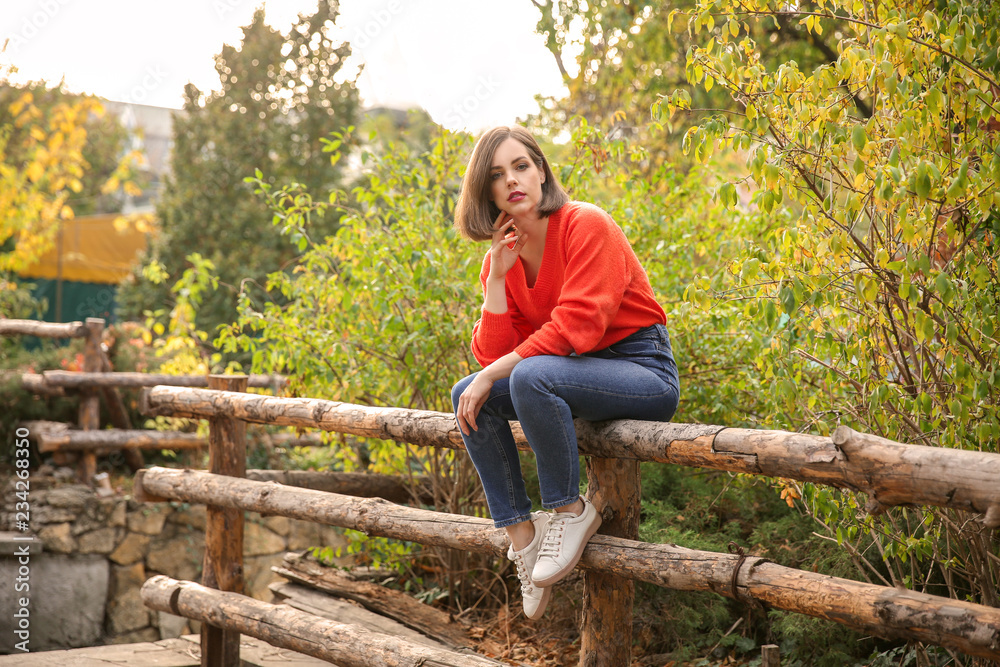 Beautiful fashionable woman sitting on wooden fence outdoors