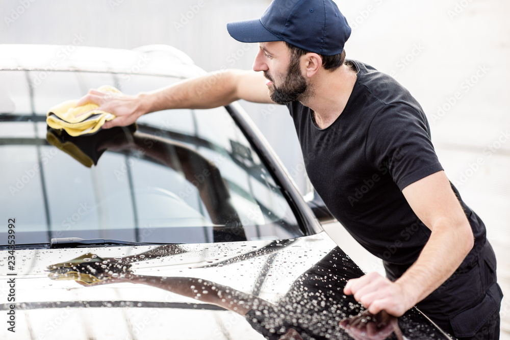 Professional washer in t-shirt and cap wiping windshield with yellow microfiber at the open air car 