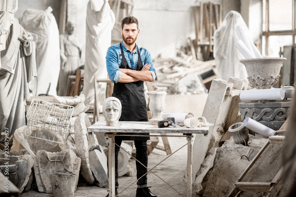 Portrait of a handsome sculptor in blue t-shirt and apron working with stone sculptures on the table