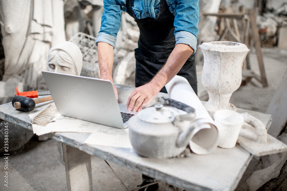 Sculptor working with laptop at the working place in the old studio with sculptures on the backgroun