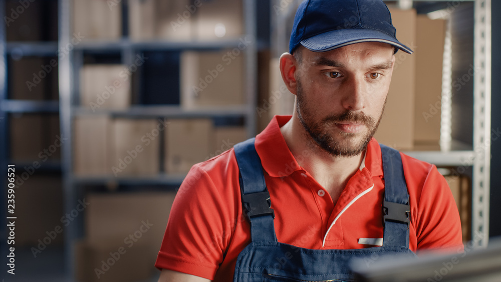 穿着制服的工人坐在仓库的桌子旁使用个人电脑的肖像。in