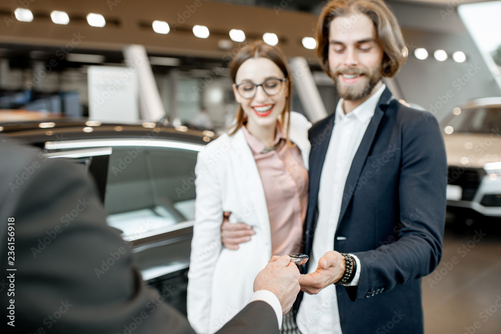 Salesperson giving keys from a new car to a lovely business couple standing together in the car show