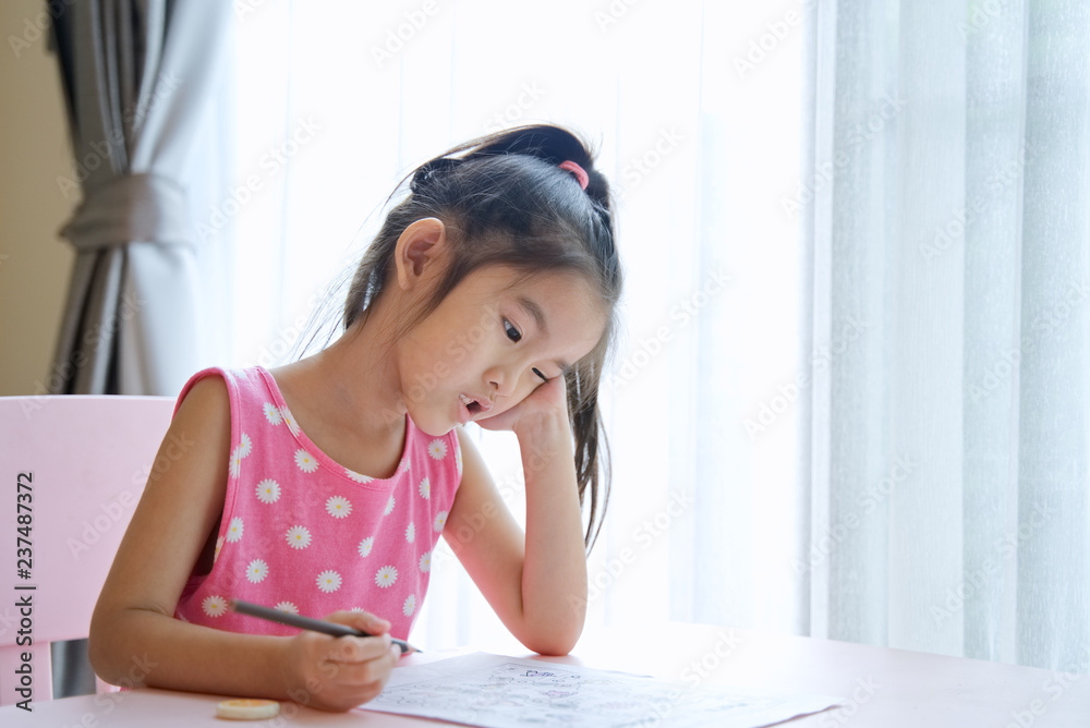 Young cute Asian kid girl is bored and tired with doing her homework on the pink table at home. It i