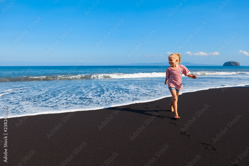 Happy barefoot kid have fun on beach walk. Run and jump by black sand along sea surf. Family travel 