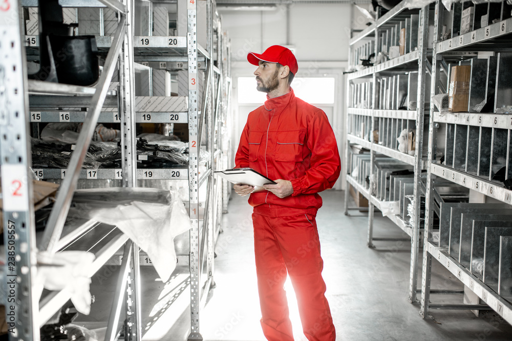 Warehouse worker in red uniform checking goods standing with clipboard in the storage with metal she