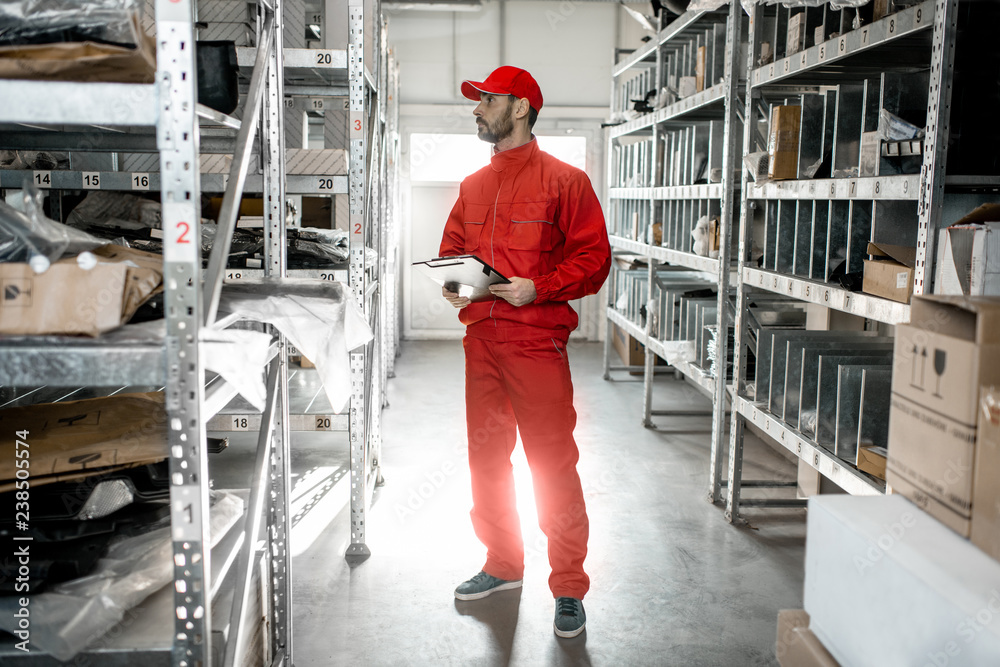 Warehouse worker in red uniform checking goods standing with clipboard in the storage with metal she