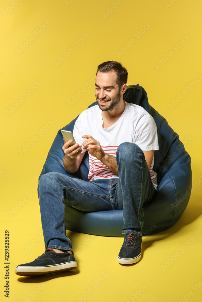 Young man with mobile phone sitting on beanbag chair against color background