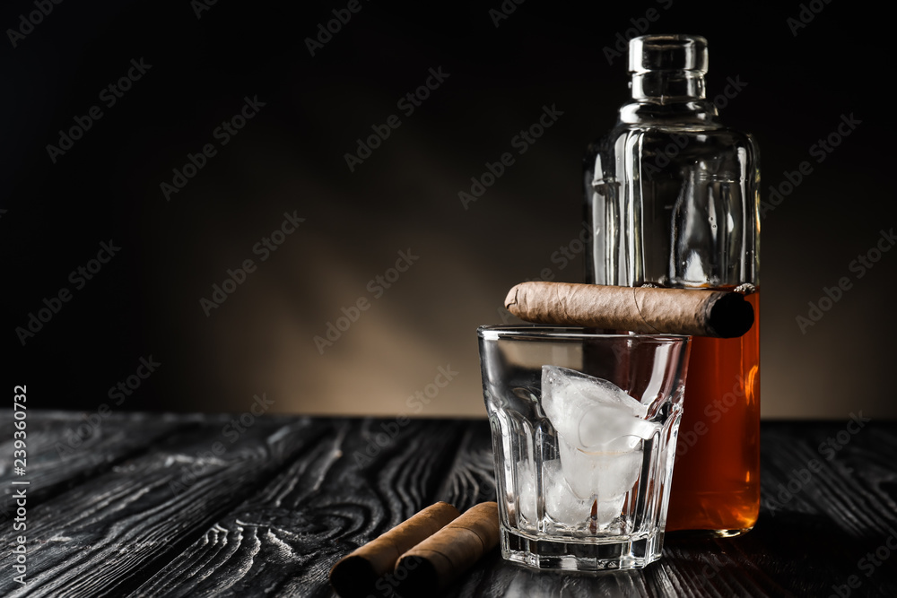 Glass with ice, bottle of whiskey and cigars on wooden table against dark background