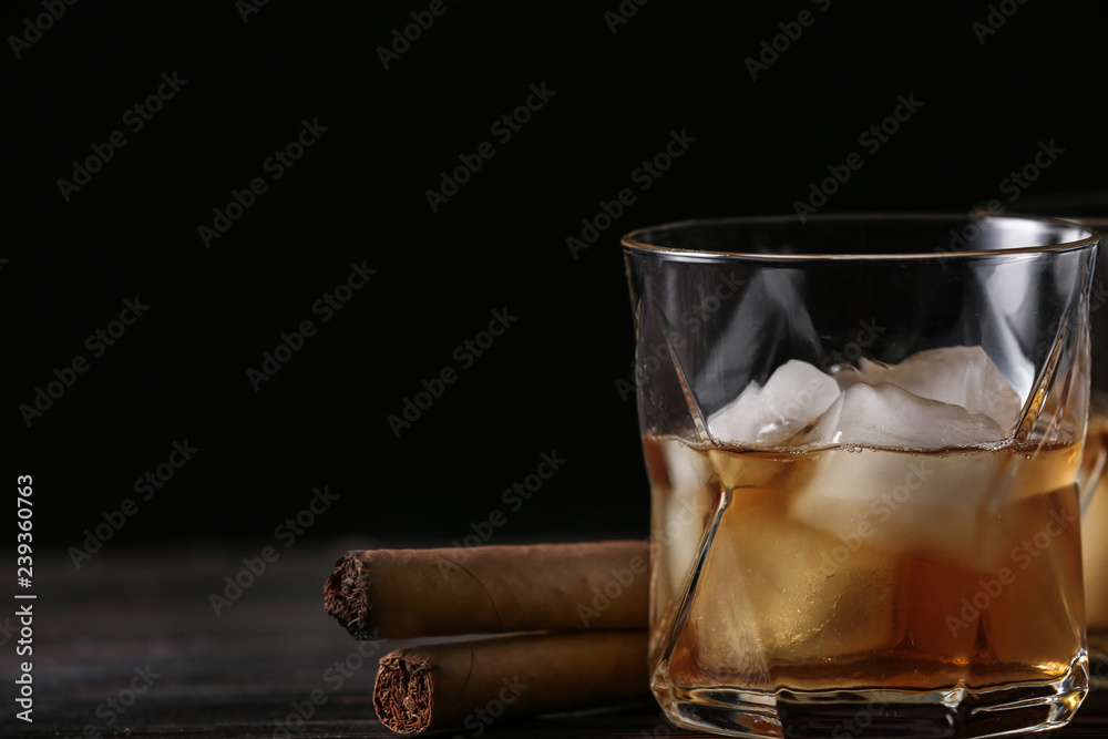 Glass of cold whiskey with cigars on wooden table against dark background