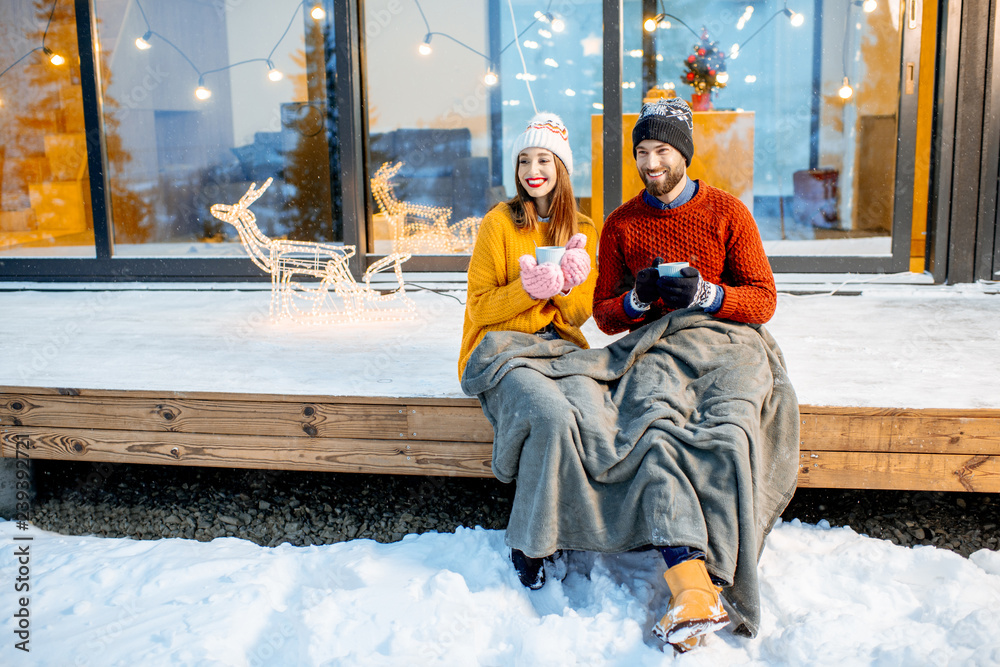 Young couple warming up with plaid and hot drinks sitting on the terrace of the modern house in the 