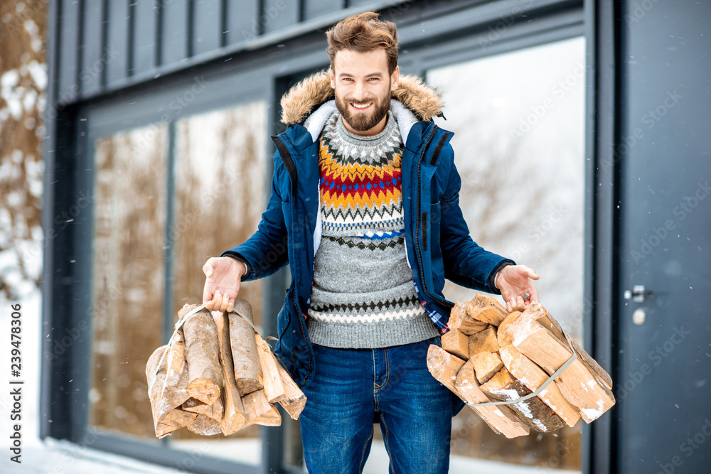 Handsome man in winter clothes carrying firewoods on the terrace near the modern house in the mounta