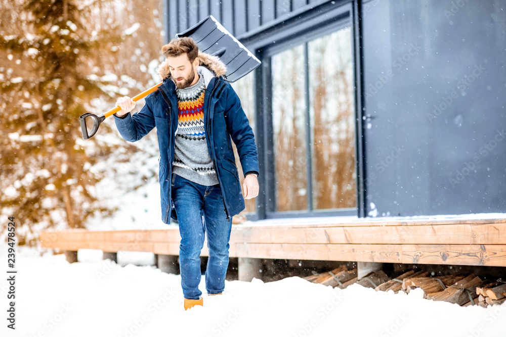 一个穿着冬装、拿着雪铲的男人在山里的现代房子附近的肖像