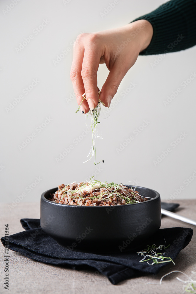 Woman adding fresh sprouts to boiled buckwheat in bowl on table