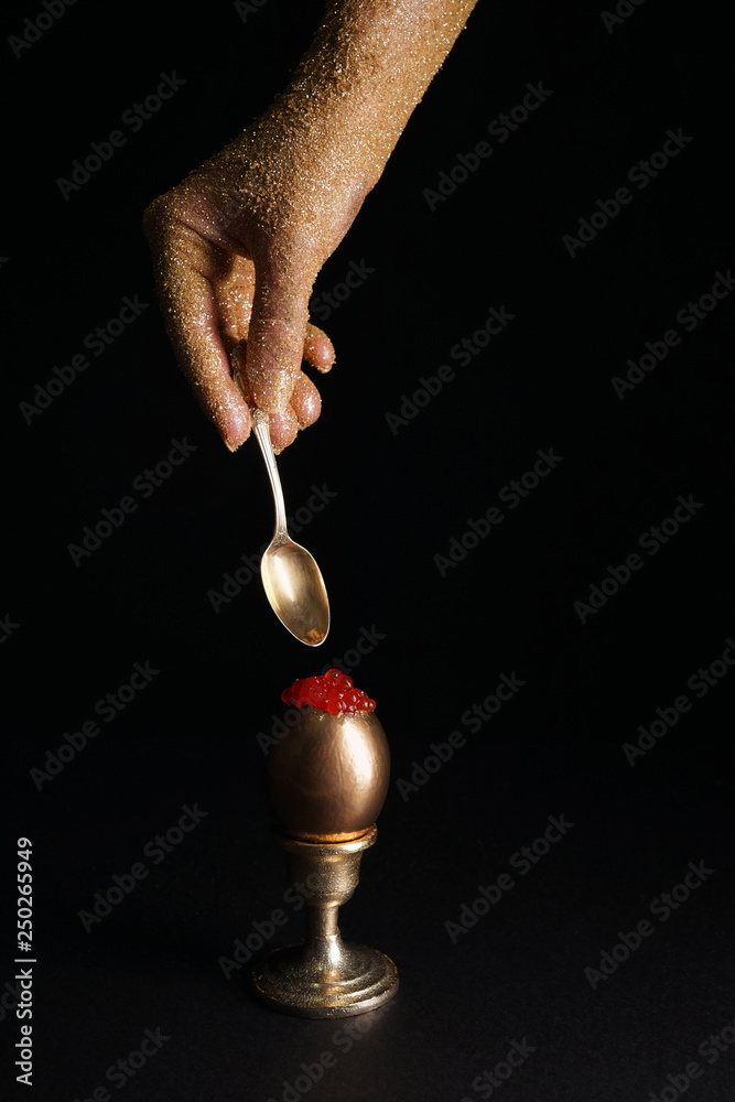 Female hand holding spoon and golden egg with red caviar on dark background