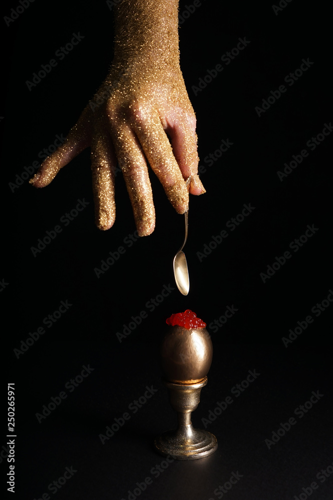 Female hand holding spoon and golden egg with red caviar on dark background