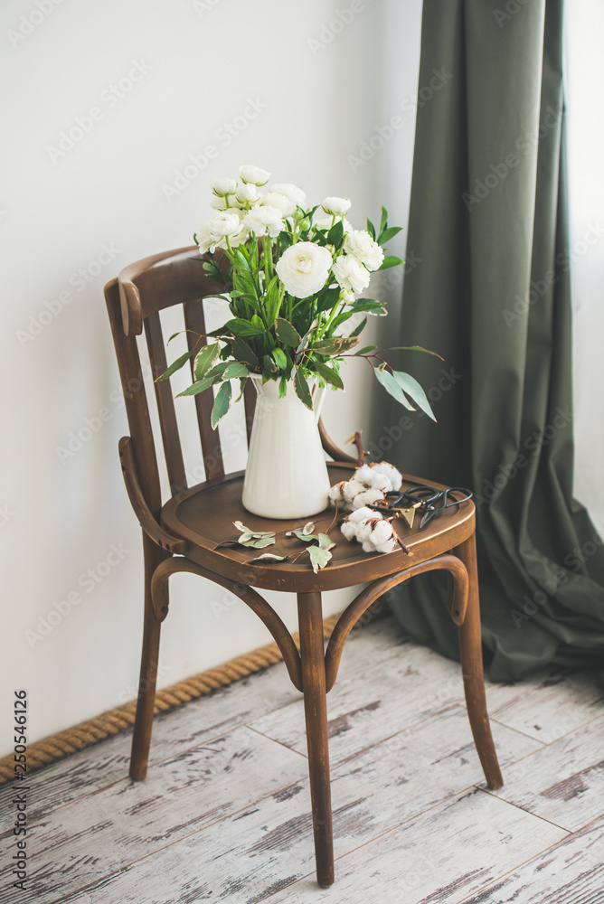 Spring white buttercup flowers in enamel jug on wooden vintage chair on floor with white wall at bac