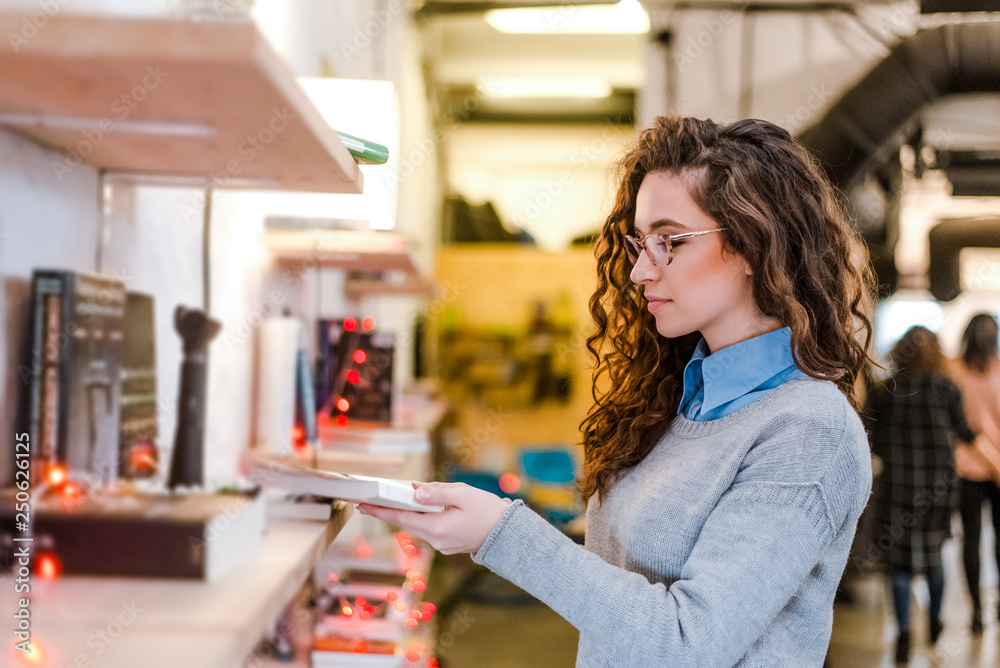 Young stylish woman choosing book from shelf.