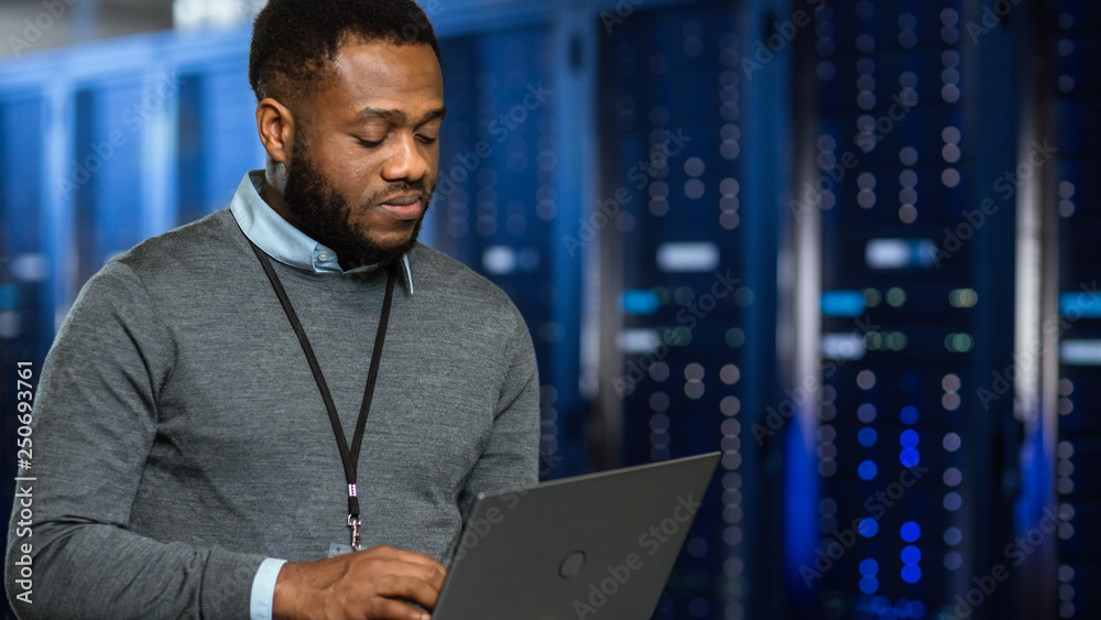 Black Data Center IT Technician Standing in Server Rack Corridor with a Laptop Computer. He is Visua