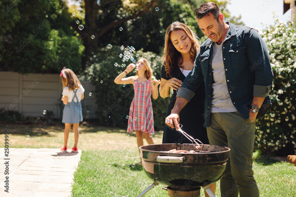 Couple cooking grilled food in backyard with children playing