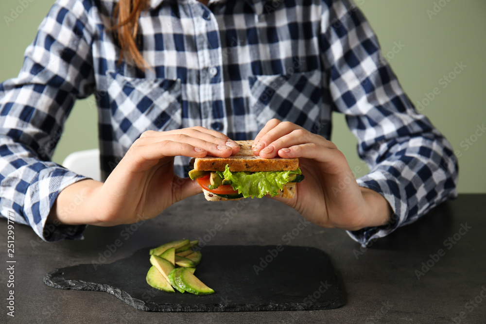 Young woman eating tasty sandwich with avocado at table, closeup