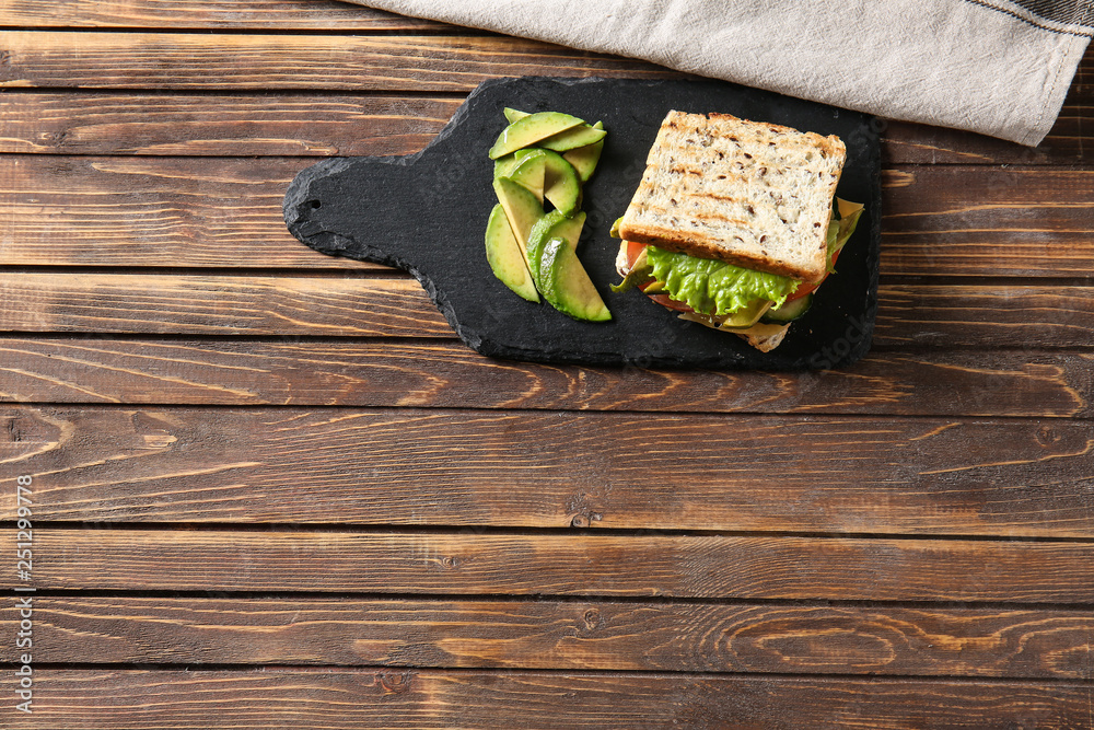 Slate plate with tasty sandwich and avocado on wooden table