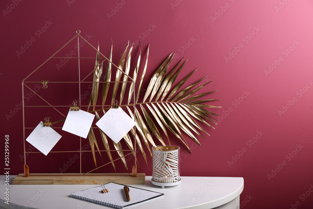 Holder with golden tropical leaf and stationery on white table near color wall