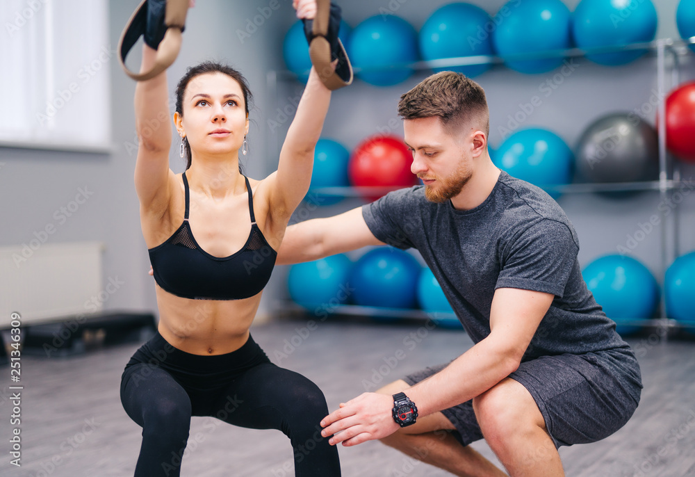 Muscular beardy trainer helping attractive female to practice with TRX in the fitness center. Sporty