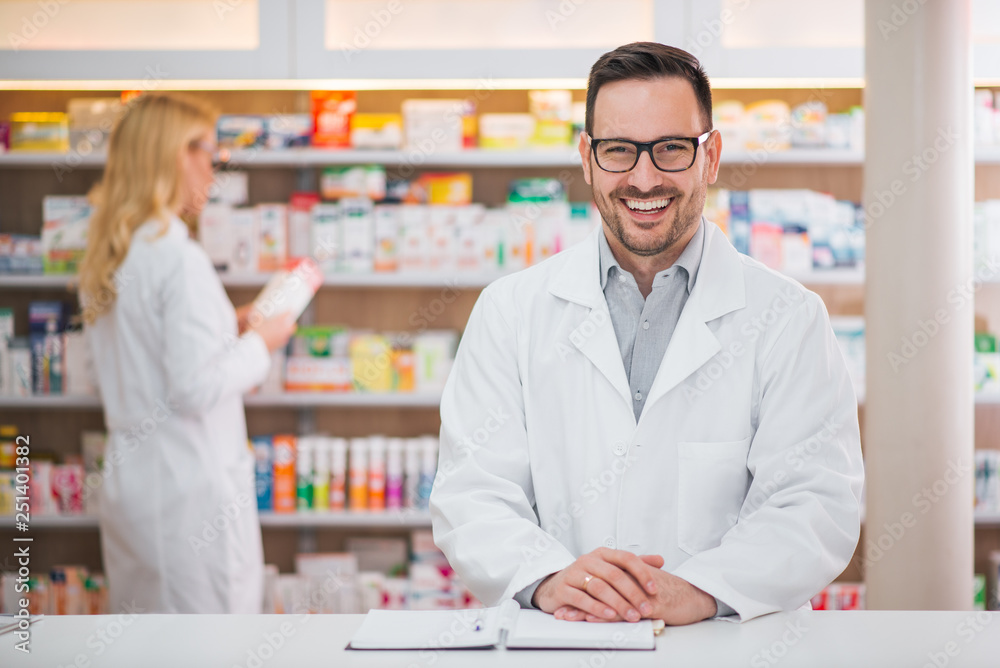 Portrait of a handsome pharmacist at the counter of a drugstore, female colleague working in the bac