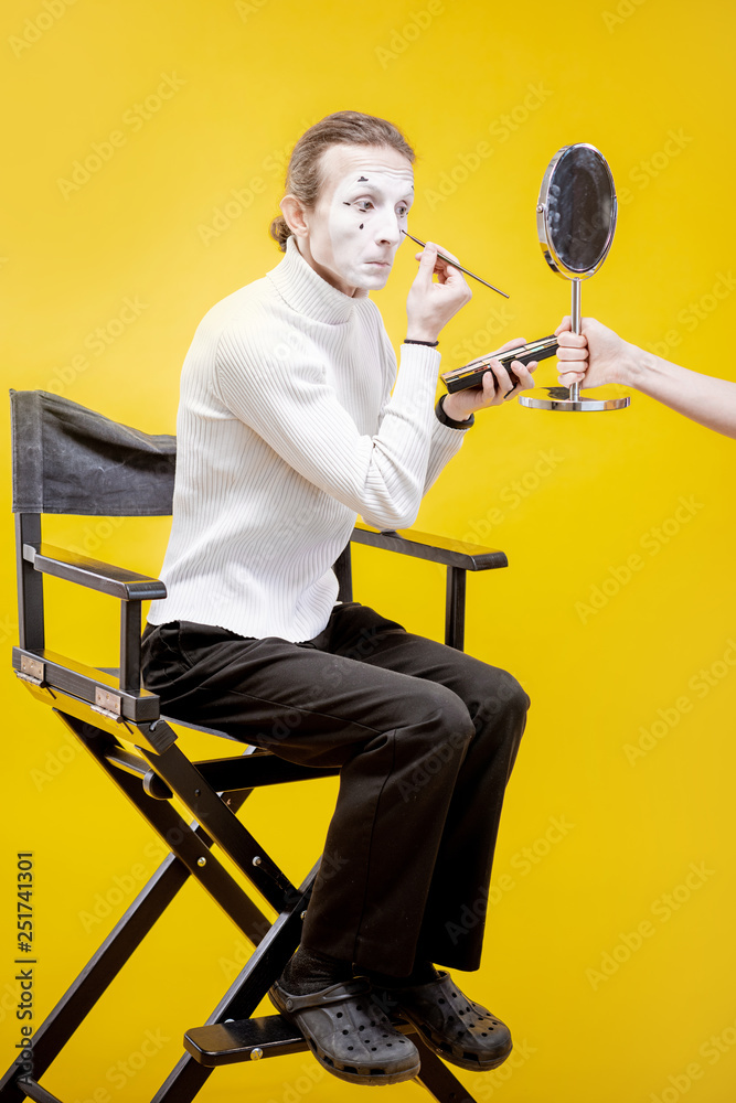 Actor applying facial makeup for pantomime performance sititng in the studio on the yellow backgroun