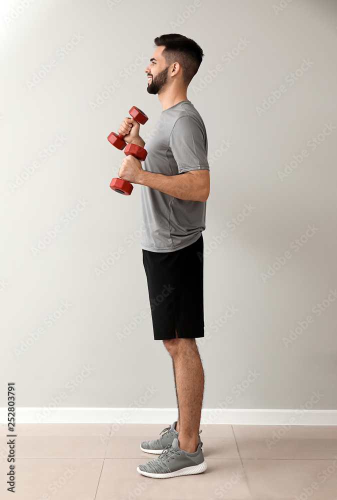 Sporty young man with dumbbells near light wall