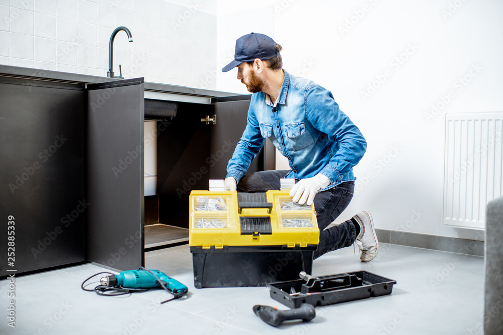 Handyman in blue shirt and cap mounting or repairing kitchen furniture in the apartment