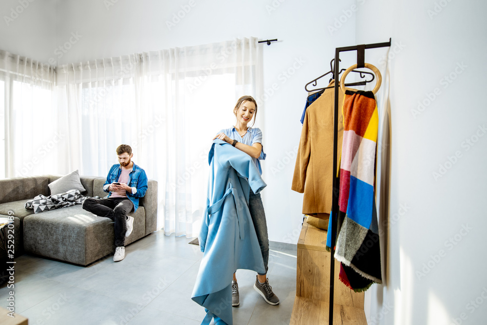 Young stylish woman wearing blue coat standing in the living room with man sitting the couch in the 