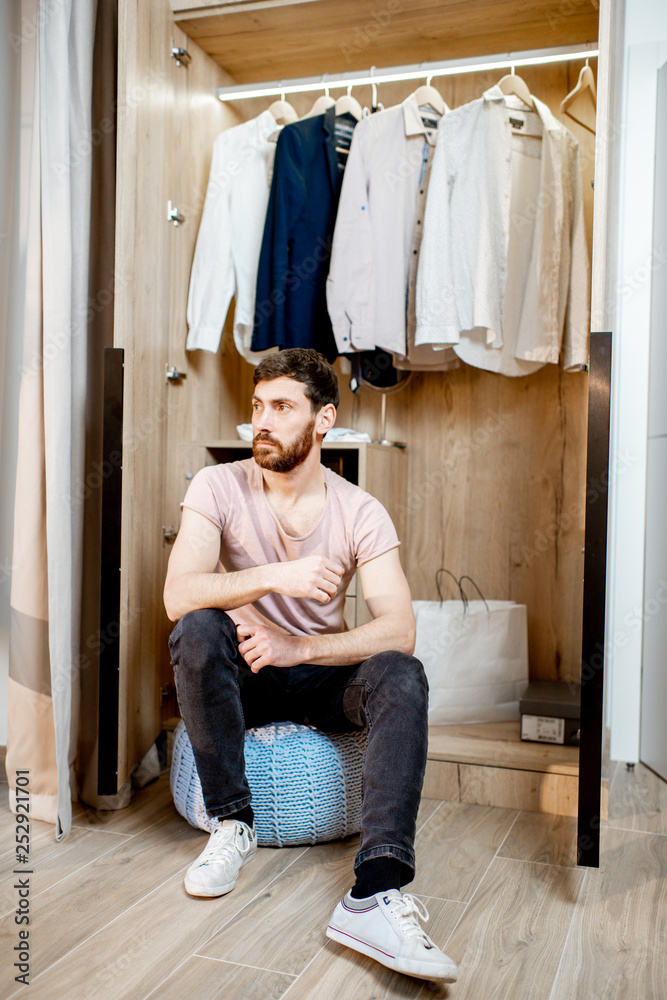 Handsome man sitting in the wardrobe full of clothes at home