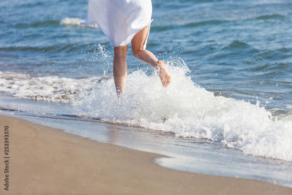 woman in white cotton dress run  through water  on sandy sea beach sunny summer day lower body back 