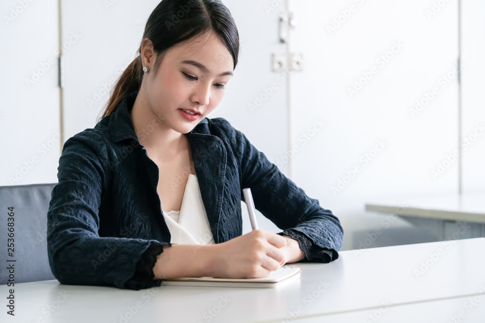 漂亮的年轻亚洲女人坐在办公桌上在笔记本上写信。内容作家a